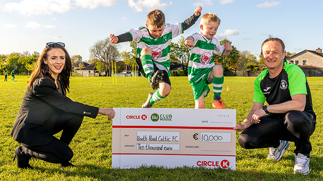 Two children in football kits joyfully jump on a grass field with two adults holding a ceremonial cheque