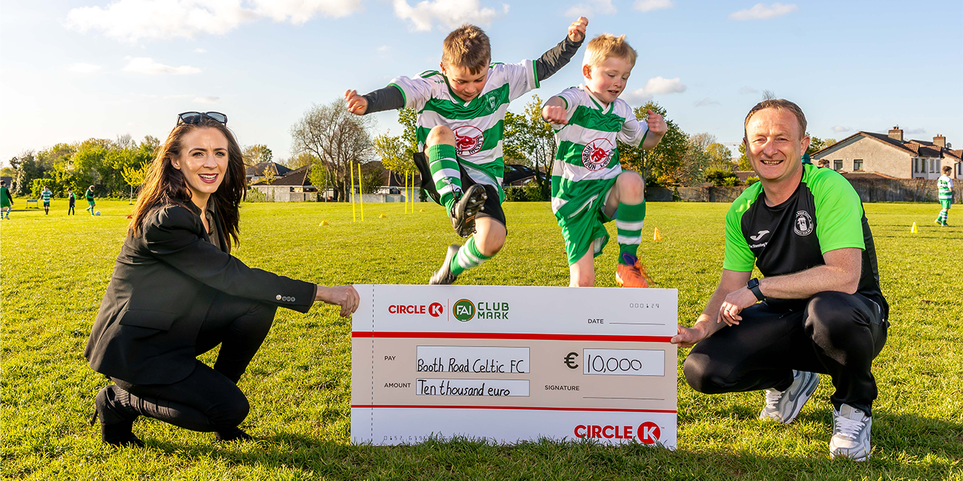 Two children in football kits joyfully jump on a grass field with two adults holding a ceremonial cheque