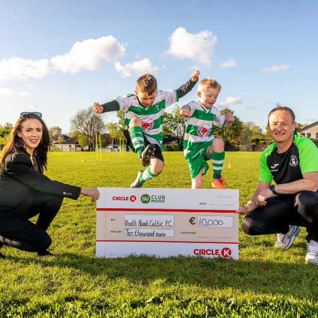 Two children in football kits joyfully jump on a grass field with two adults holding a ceremonial cheque