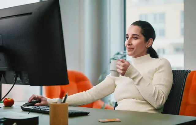 Woman in an office happily working on her computer, with coffee in hand