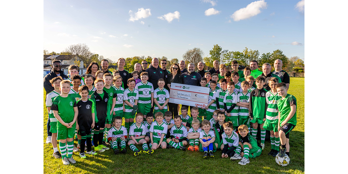 Large group of children and adults posing outdoors on a football pitch, holding a ceremonial cheque